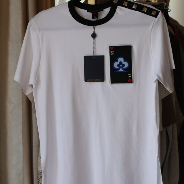 Biały t-shirt Louis Vuitton z czarną lamówką przy dekolcie, naszywką w formie karty do gry na piersi i metalowymi ćwiekami na lewym ramieniu, wiszący na złotym wieszaku wraz z metkami