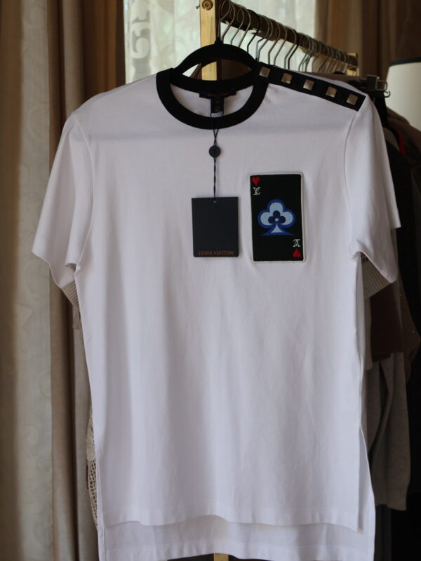 Biały t-shirt Louis Vuitton z czarną lamówką przy dekolcie, naszywką w formie karty do gry na piersi i metalowymi ćwiekami na lewym ramieniu, wiszący na złotym wieszaku wraz z metkami