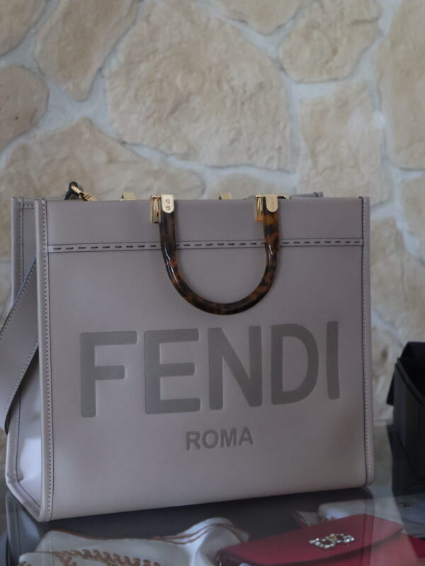 Szaro-beżowa skórzana torebka typu shopper Fendi Sunshine z wielkim wytłoczonym logo i sztywnymi rączkami z efektem szylkretu, stojąca na tle kamiennej ściany