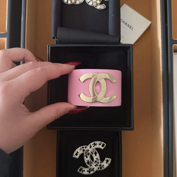 Szeroka różowa bransoletka Chanel z dużym złotym logo CC, trzymana w dłoni nad oryginalnym czarnym pudełkiem marki