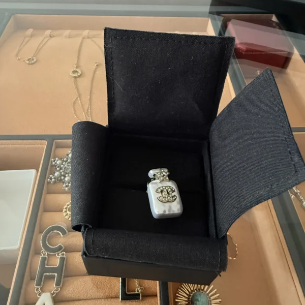 Broszka Chanel w kształcie małego, białego flakonu perfum ze lśniącym logo CC i gwiazdką na zatyczce, umieszczona w otwartym, czarnym pudełku marki