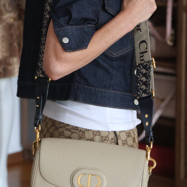 Beżowa torebka Dior Bobby Medium ze złotym logo CD, noszona na szerokim pasku Dior Oblique przez modelkę w jeansowej kurtce i spodniach w monogram.
