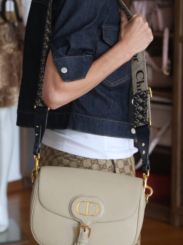 Beżowa torebka Dior Bobby Medium ze złotym logo CD, noszona na szerokim pasku Dior Oblique przez modelkę w jeansowej kurtce i spodniach w monogram.