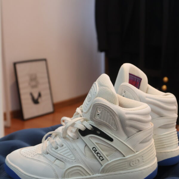 Białe wysokie sneakersy Gucci z logo na bocznym pasku, sznurowaniem, perforowanym noskiem i niebieską podeszwą, ustawione na granatowym pikowanym pufie we wnętrzu.
