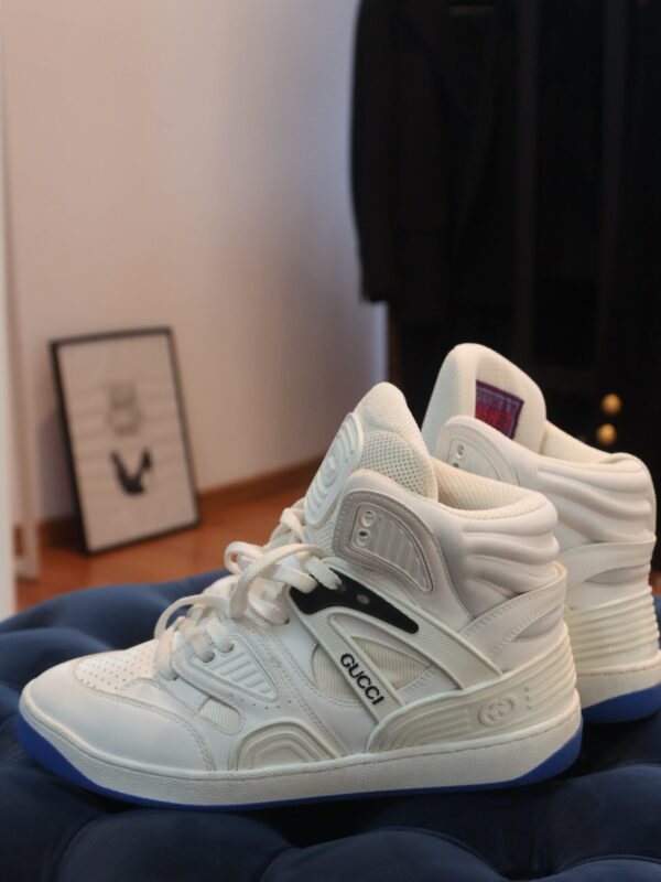 Białe wysokie sneakersy Gucci z logo na bocznym pasku, sznurowaniem, perforowanym noskiem i niebieską podeszwą, ustawione na granatowym pikowanym pufie we wnętrzu.