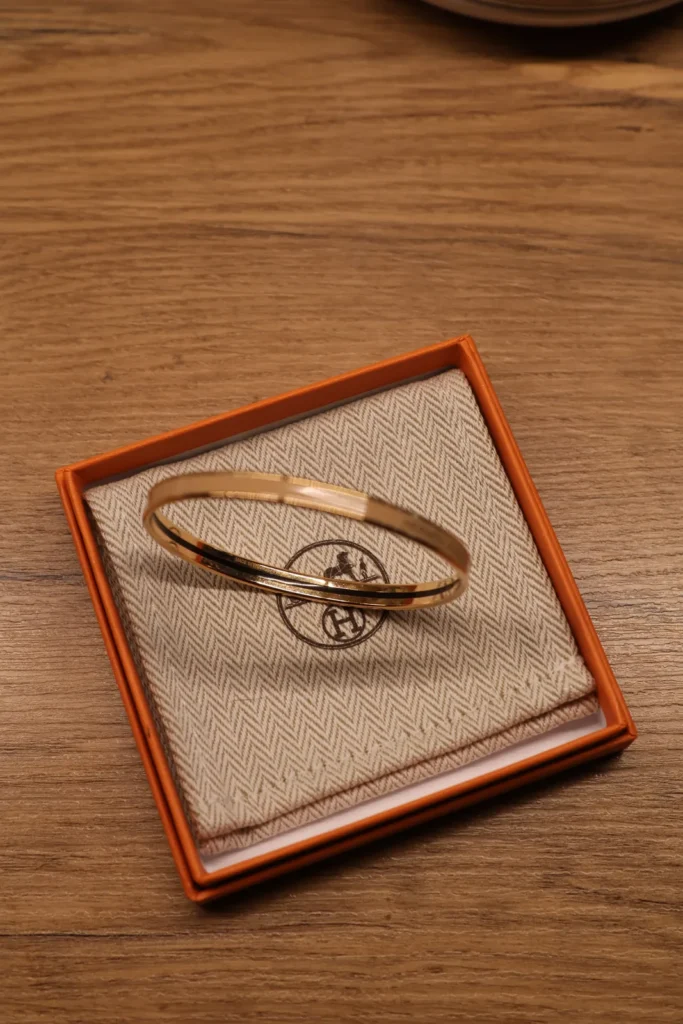 Bransoletka Hermès w kolorze złotym na dustbagu w pomarańczowym pudełku Hermès