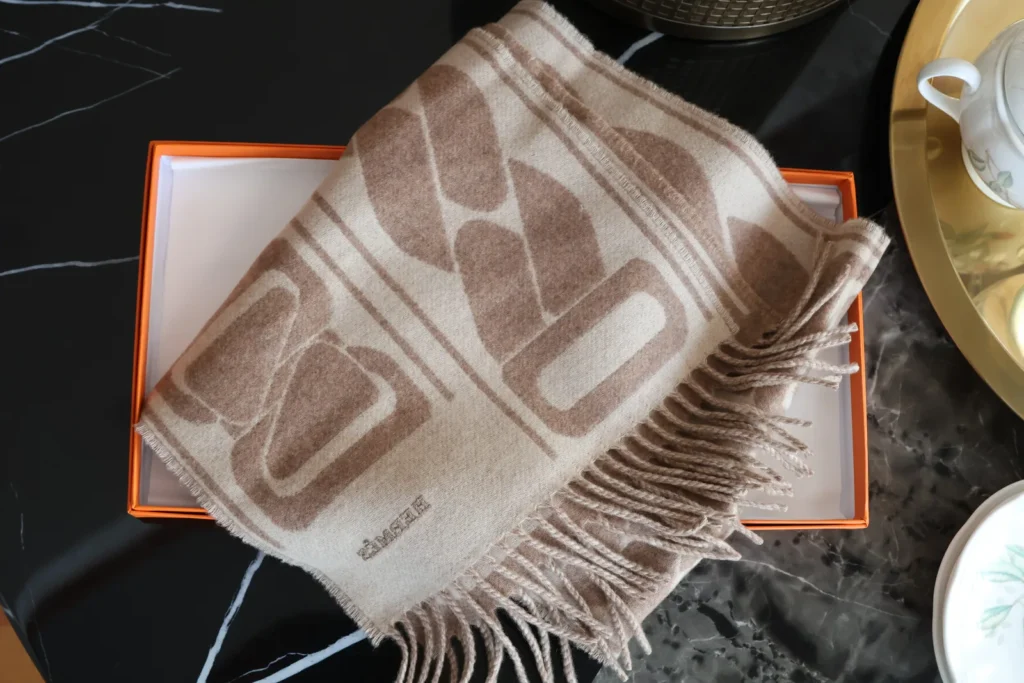 Beżowy szalik Hermès z kaszmiru z dużym wzorem w odcieniach beżu i brązu, z frędzlami, ułożony na pomarańczowym pudełku Hermès.