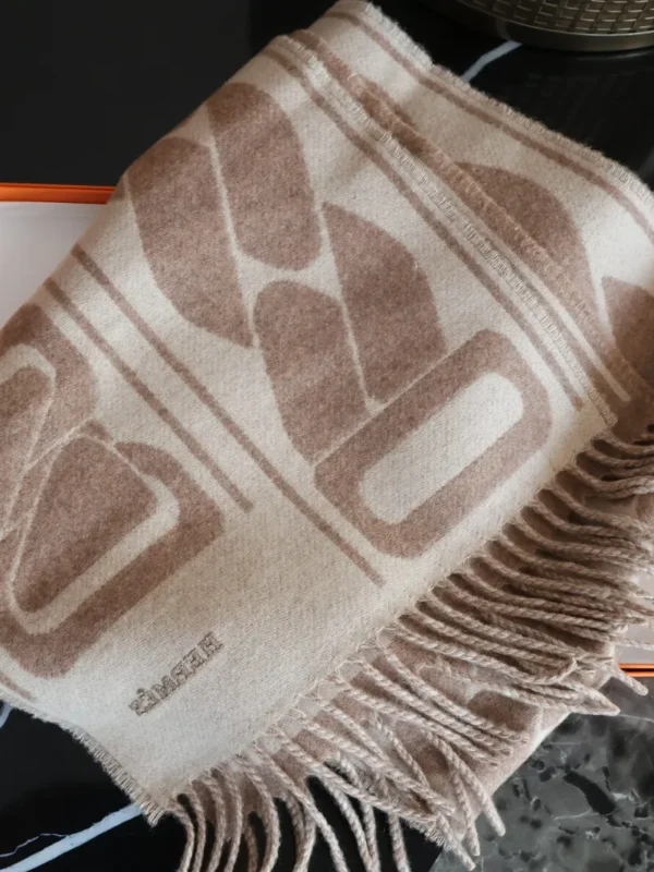 Beżowy szalik Hermès z kaszmiru z dużym wzorem w odcieniach beżu i brązu, z frędzlami, ułożony na pomarańczowym pudełku Hermès.