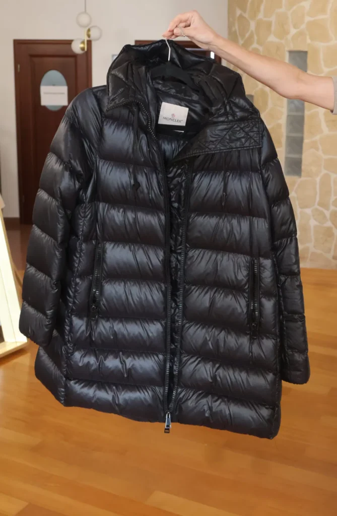 Czarna pikowana kurtka Moncler z połyskiem i kapturem, zapinana na zamek, na wieszaku.