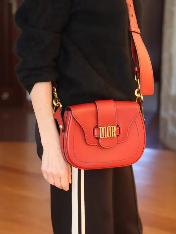 Czerwona torebka Dior z zaokrągloną klapą i złotym logo DIOR, noszona na szerokim pasku na ramię.