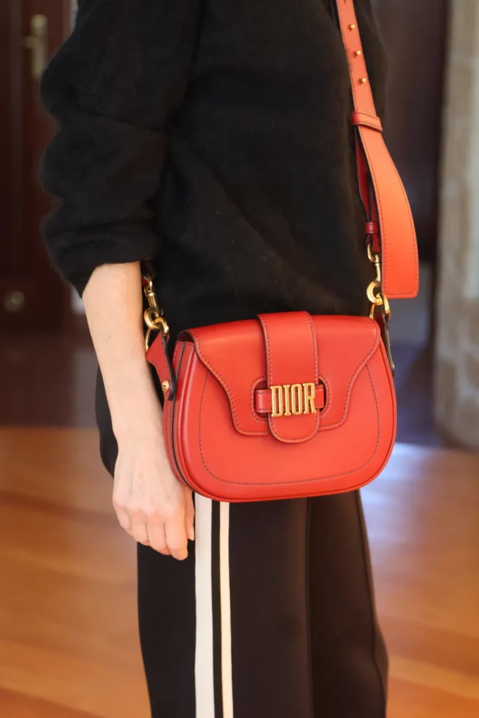 Czerwona torebka Dior z zaokrągloną klapą i złotym logo DIOR, noszona na szerokim pasku na ramię.